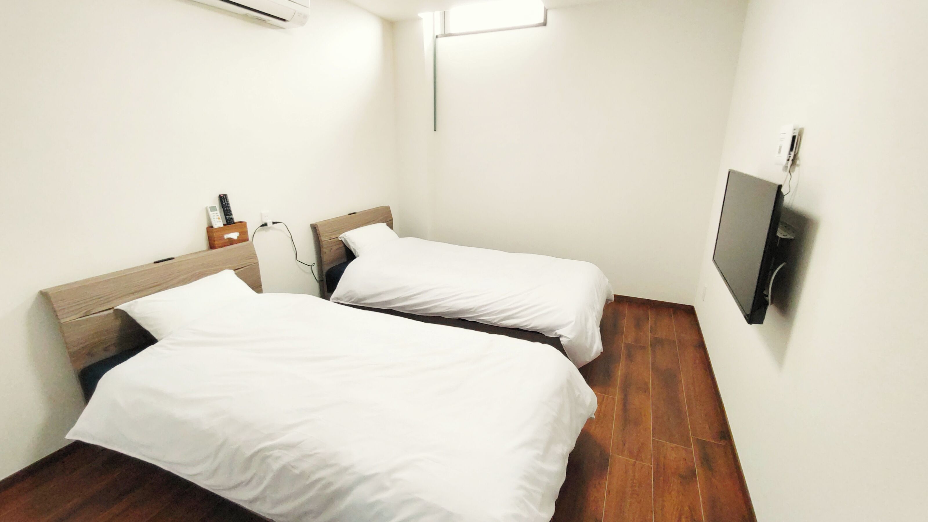 A small, clean room with two single beds, a TV mounted on the wall, and a wooden nightstand with a phone and remote.