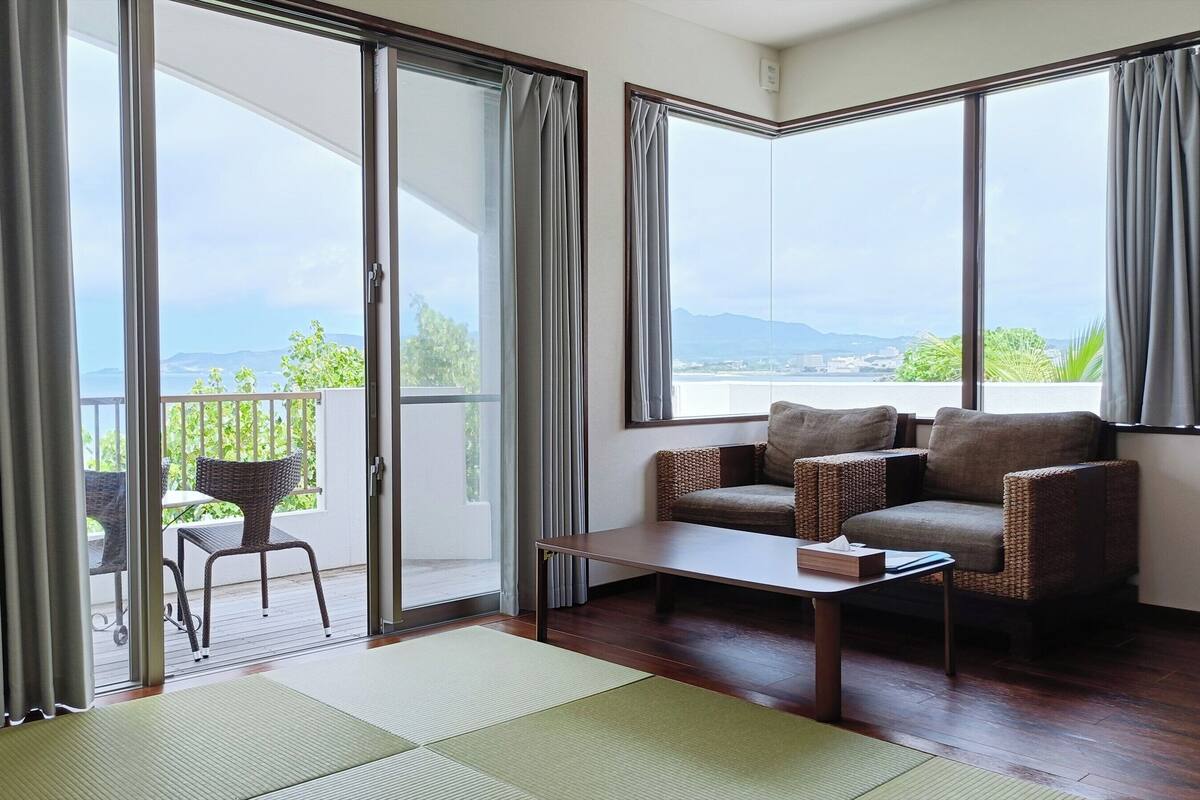 Una habitación con puerta corrediza de vidrio que da a un balcón, un sofá, una mesita y vistas a zonas verdes y montañas.
