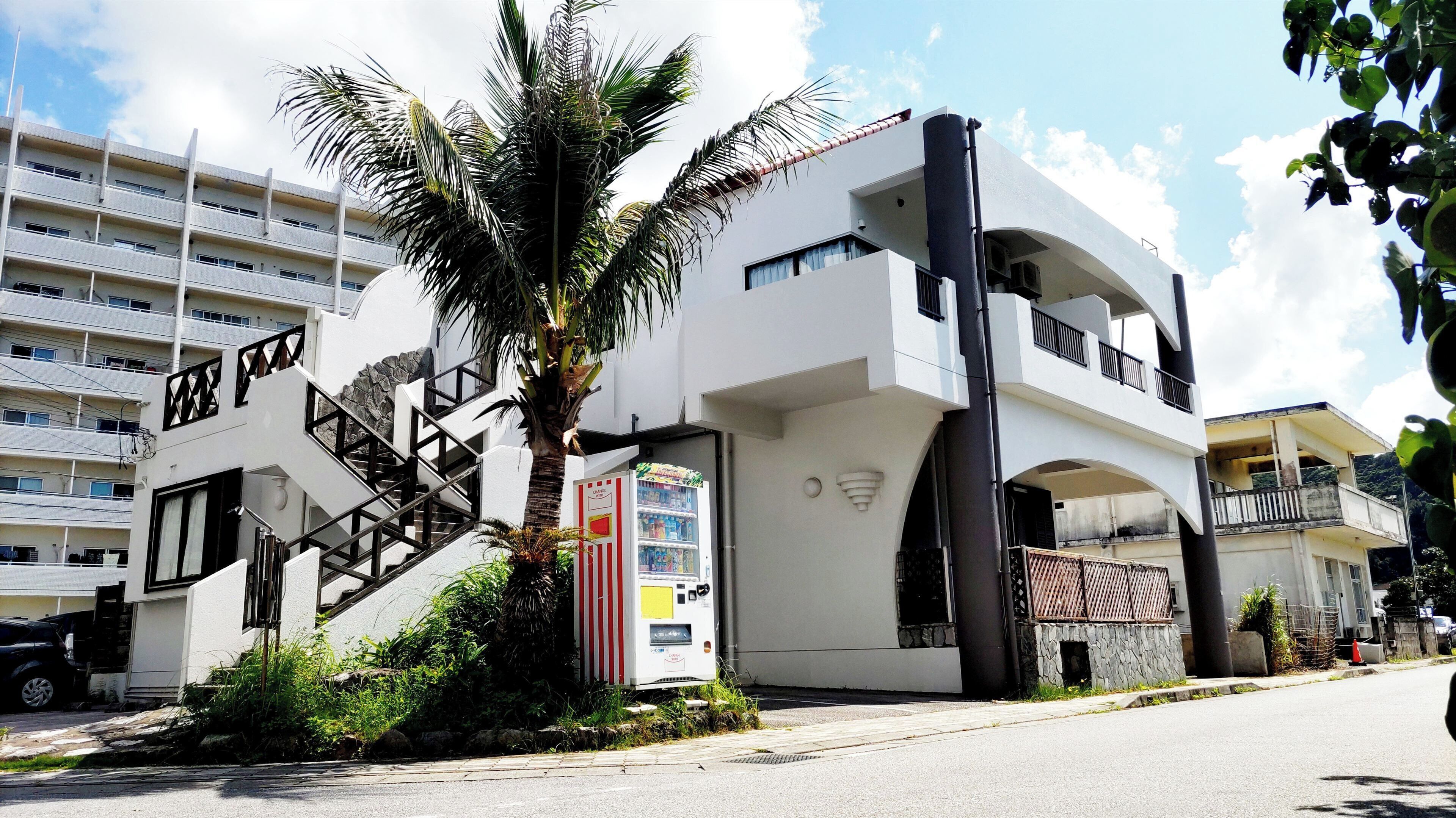 A modern building with a white facade, a staircase leading to a balcony, and a palm tree in the foreground.