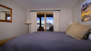 A bedroom with a large bed, a window with curtains, a chair, and a painting on the wall. - Ideal Mountain Condo! Ski-In/Ski-Out, Slopeside with Beautiful Views (Mt. Crested Butte)