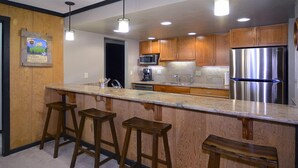 A modern kitchen with wooden cabinets, a stainless steel refrigerator, and a granite countertop with bar stools. - Ideal Mountain Condo! Ski-In/Ski-Out, Slopeside with Beautiful Views (Mt. Crested Butte)