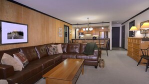 A modern living room with a brown leather sofa, a wooden coffee table, and a kitchen in the background. - Ideal Mountain Condo! Ski-In/Ski-Out, Slopeside with Beautiful Views (Mt. Crested Butte)