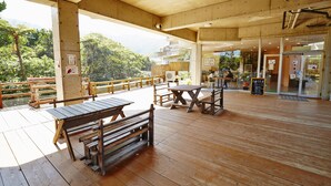 Une terrasse extérieure en bois, dotée de tables et de bancs, un espace couvert offrant une vue sur une montagne boisée, et un panneau indiquant la présence d’un café.