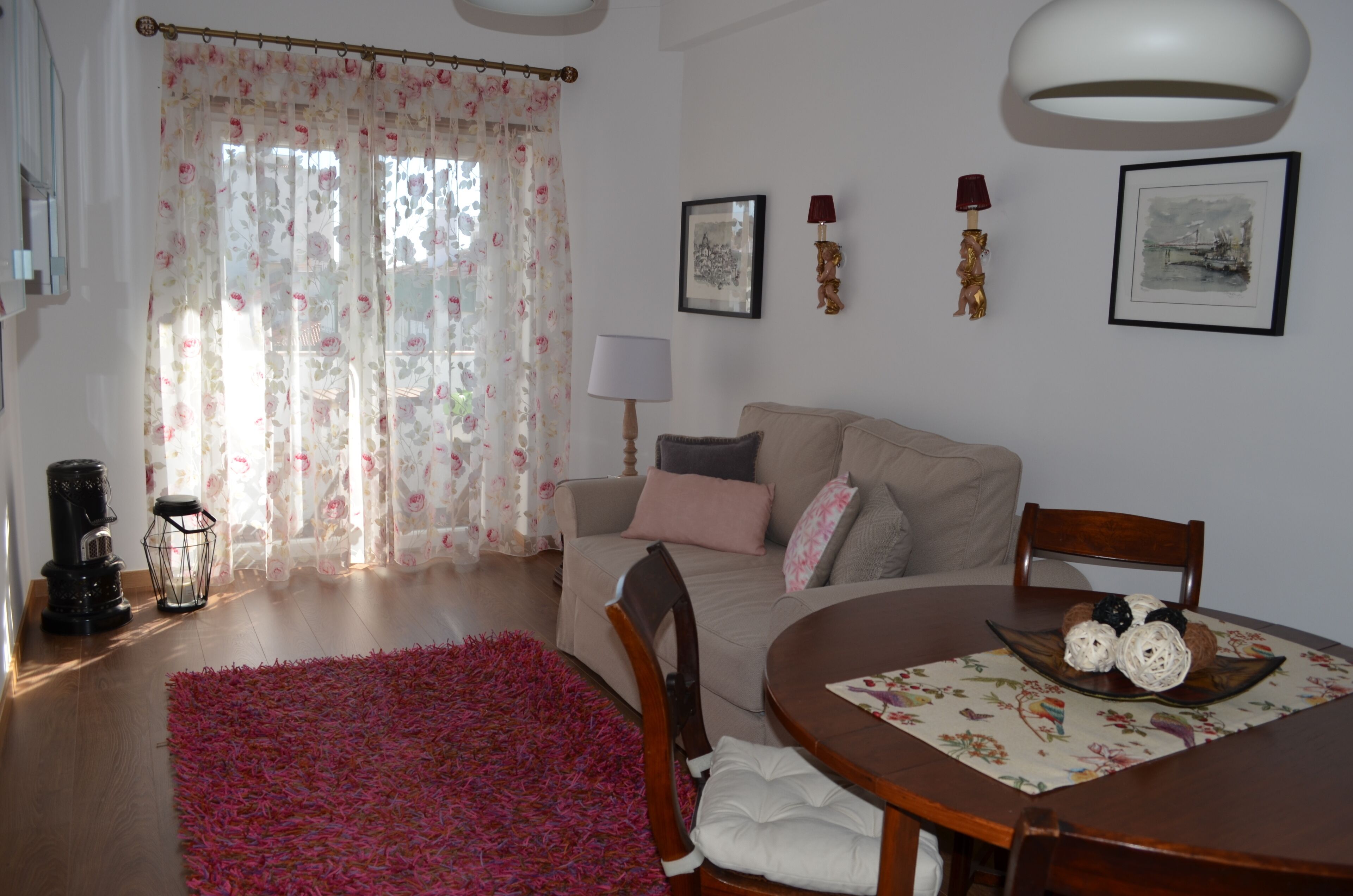 A living room with a sofa, a dining table with a placemat, a coffee table, a pink rug, a lamp, and a picture frame on the wall.