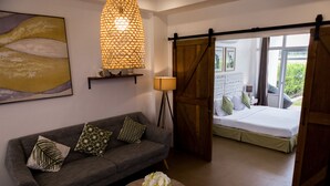 A modern living room with a grey sofa, a round coffee table with a vase, a large abstract painting, a hanging light fixture, and a sliding barn door leading to a bedroom with a bed and a view of greenery. - The Mansion (Mabalacat City)