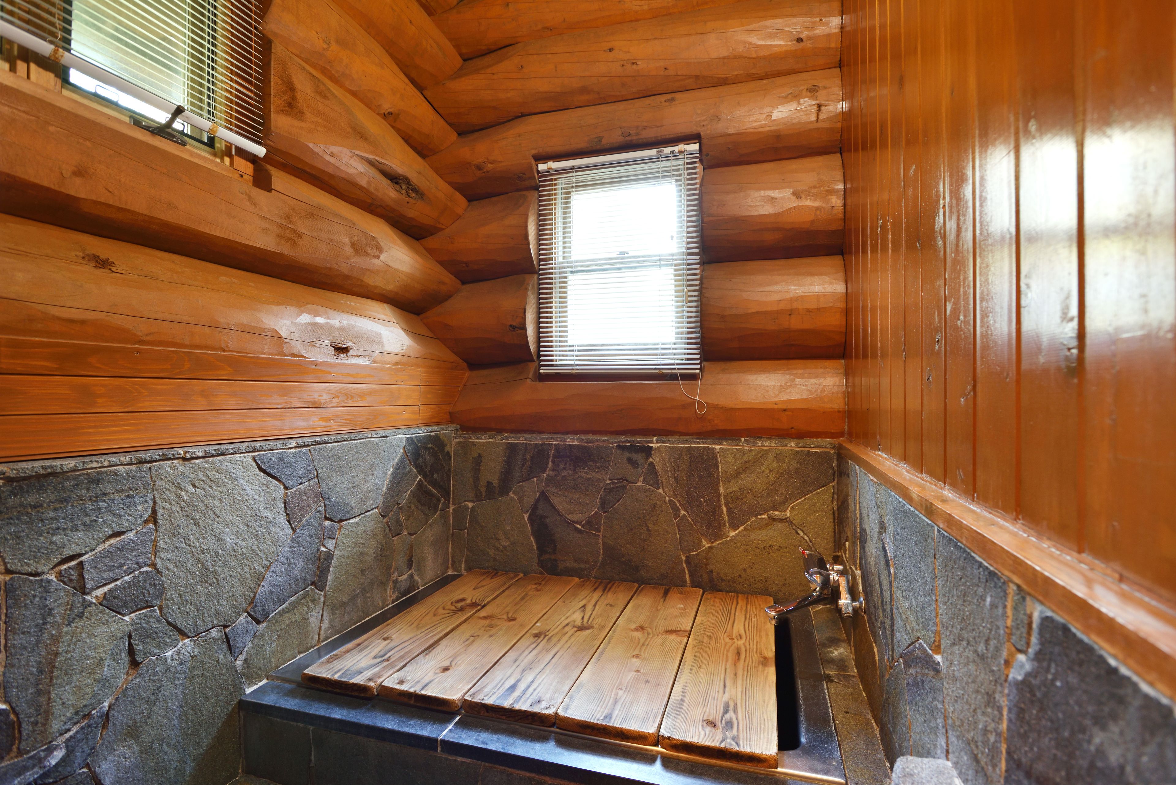 A log cabin interior with a stone wall, wooden floor, and a window with blinds.
