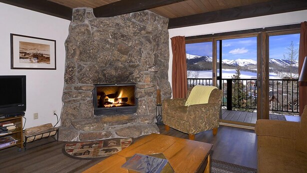 A cozy living room with a stone fireplace, a television, and a view of mountains through large windows. - Great Location! Ski-In/Out Condo. Mountain Views, Fireplace. (Mt. Crested Butte)