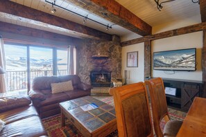 Een gezellige woonkamer met een stenen openhaard, leren banken en een groot raam met uitzicht op een besneeuwd berglandschap.