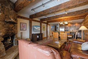 A cozy living room with a fireplace, leather sofas, wooden beams, and a kitchen in the background.