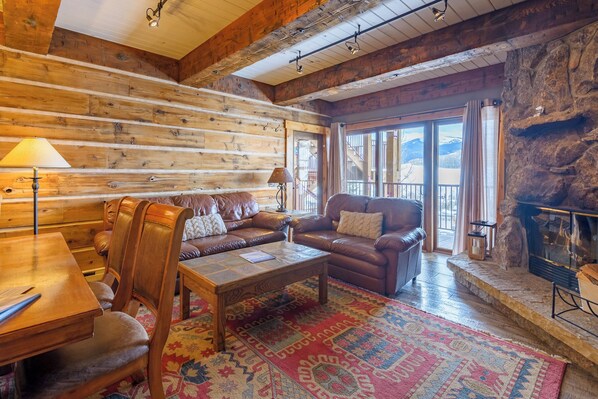 A log cabin interior with a wooden dining table, leather sofas, a patterned rug, a fireplace, and a large window with a mountain view.