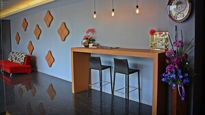 A reception area with a wooden desk, two black barstools, a red sofa, decorative wall panels, hanging pendant lights, and a clock on the wall. - Merdelong Hotel (Phatthalung)
