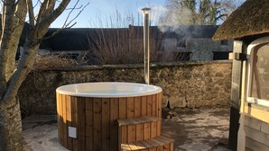 A wooden hot tub with a chimney, surrounded by stone walls and a tree.