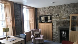 Una habitación acogedora con pared de piedra, chimenea, un sillón a cuadros, sofá rojo, mesa de madera con lámpara y televisor sobre un mueble de madera.
