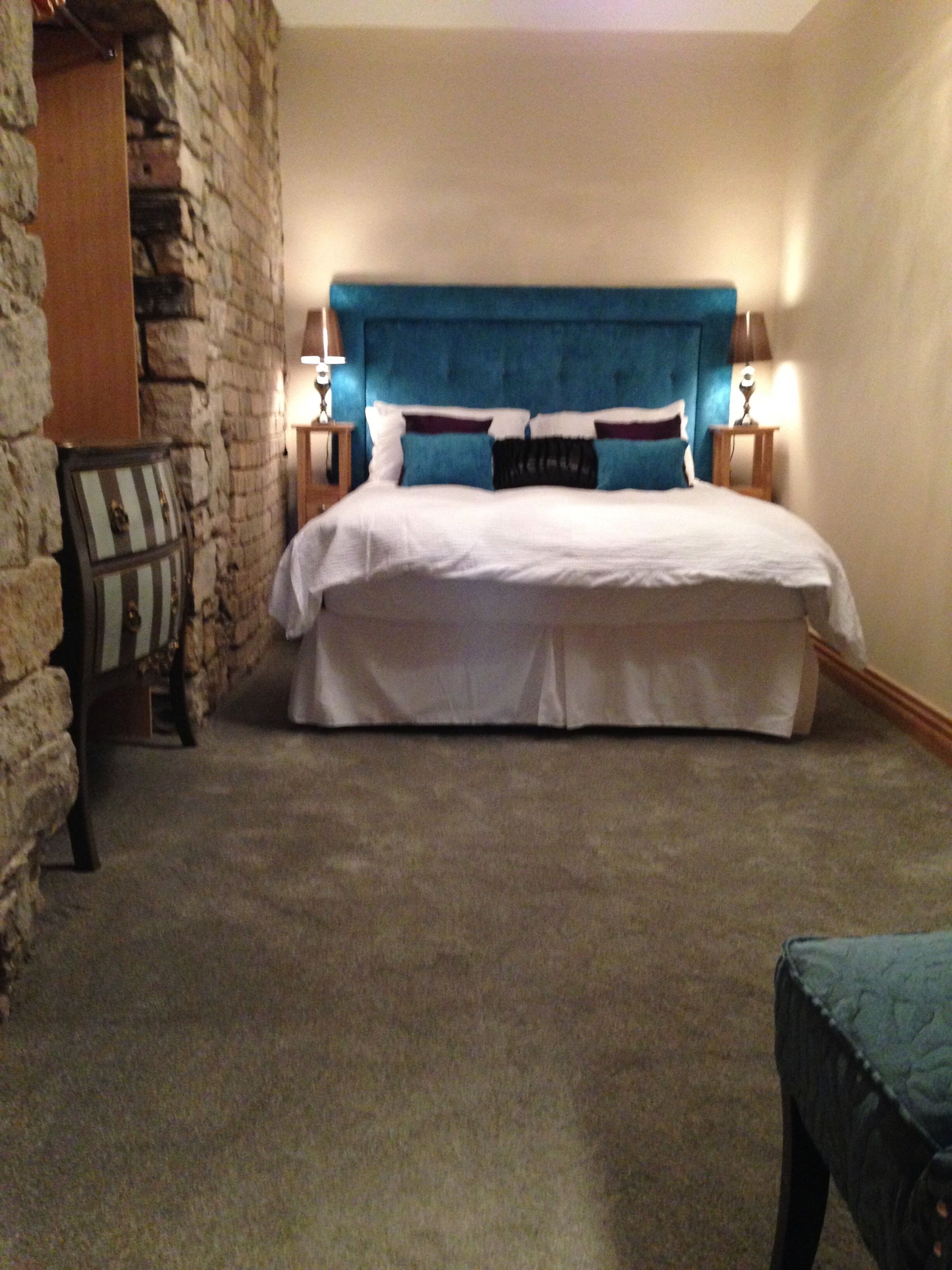 Un dormitorio con pared de piedra, una cama con cabecera azul, dos mesitas de noche con lámparas y una maleta apoyada contra la pared.