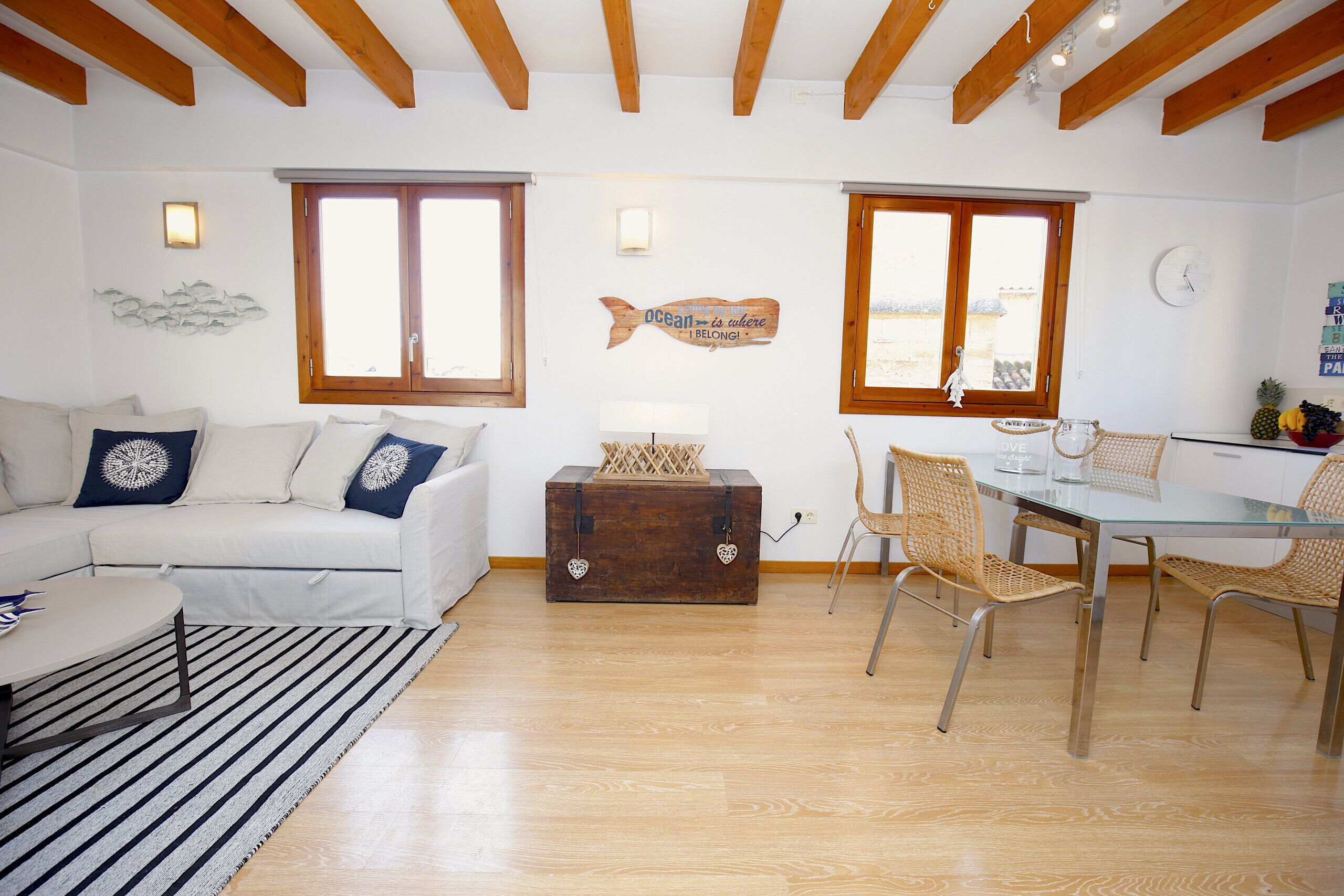 A living room with a white sofa, a glass dining table, wicker chairs, wooden flooring, and a wooden ceiling.