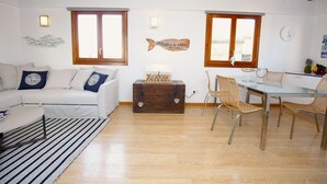 Ein Wohnzimmer mit weißem Sofa, einem Glastisch, Korbstühlen, Holzboden und einer Holzbalkendecke.