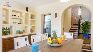 Un coin repas avec une table en bois, un panier de fruits et une bougie bleue. On aperçoit, au fond, une cuisine avec un réfrigérateur et des placards.