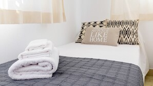 Un lit bien fait, avec une serviette blanche pliée, une couverture grise à texture intéressante et deux coussins décoratifs à motifs géométriques, l’un d’eux portant une citation inspirante.