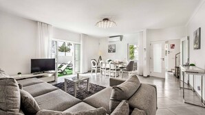 Ein modernes Wohnzimmer mit einer grauen Eckcouch, einem Flachbildfernseher und einem Essbereich mit weißen Stühlen und einem Tisch.