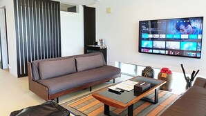 A modern living room with a sofa, coffee table, and a flat-screen TV displaying a video call interface. - Kariyushi Condominium Resort Onna Maeda Base (Onna)