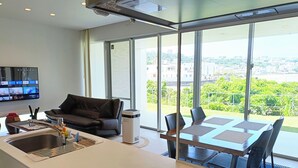 A modern living room with a sofa, a dining table with chairs, a large window with a city view, and a flat-screen TV mounted on the wall. - Kariyushi Condominium Resort Onna Maeda Base (Onna)