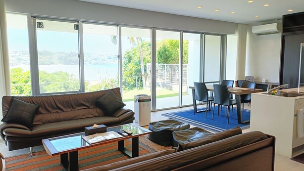 A modern living room with a brown leather sofa, a glass coffee table, and a dining area with a wooden table and chairs. - Kariyushi Condominium Resort Onna Maeda Base (Onna)