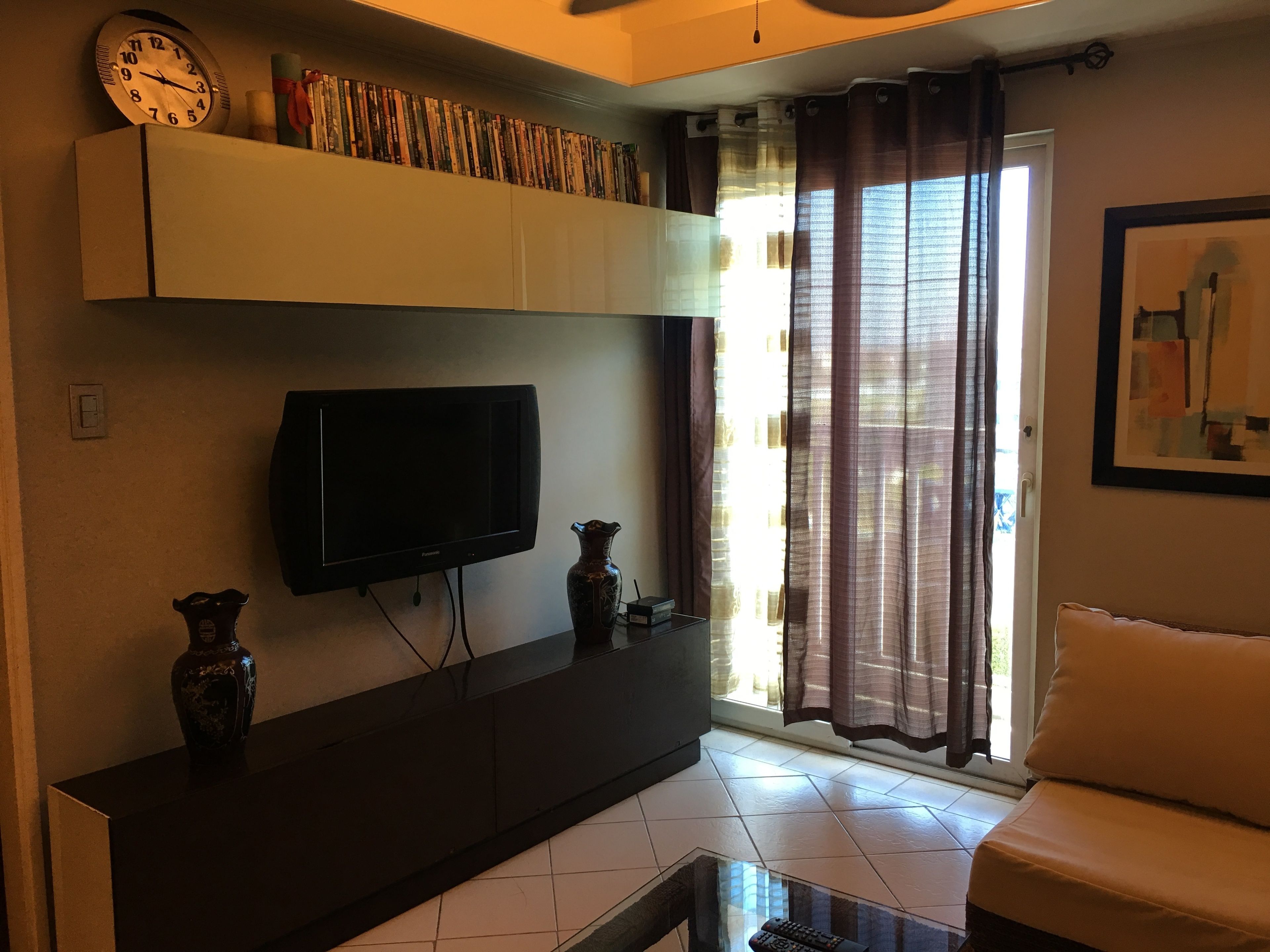 A living room with a flat-screen TV, a bookshelf with books, a vase, and a window with curtains.