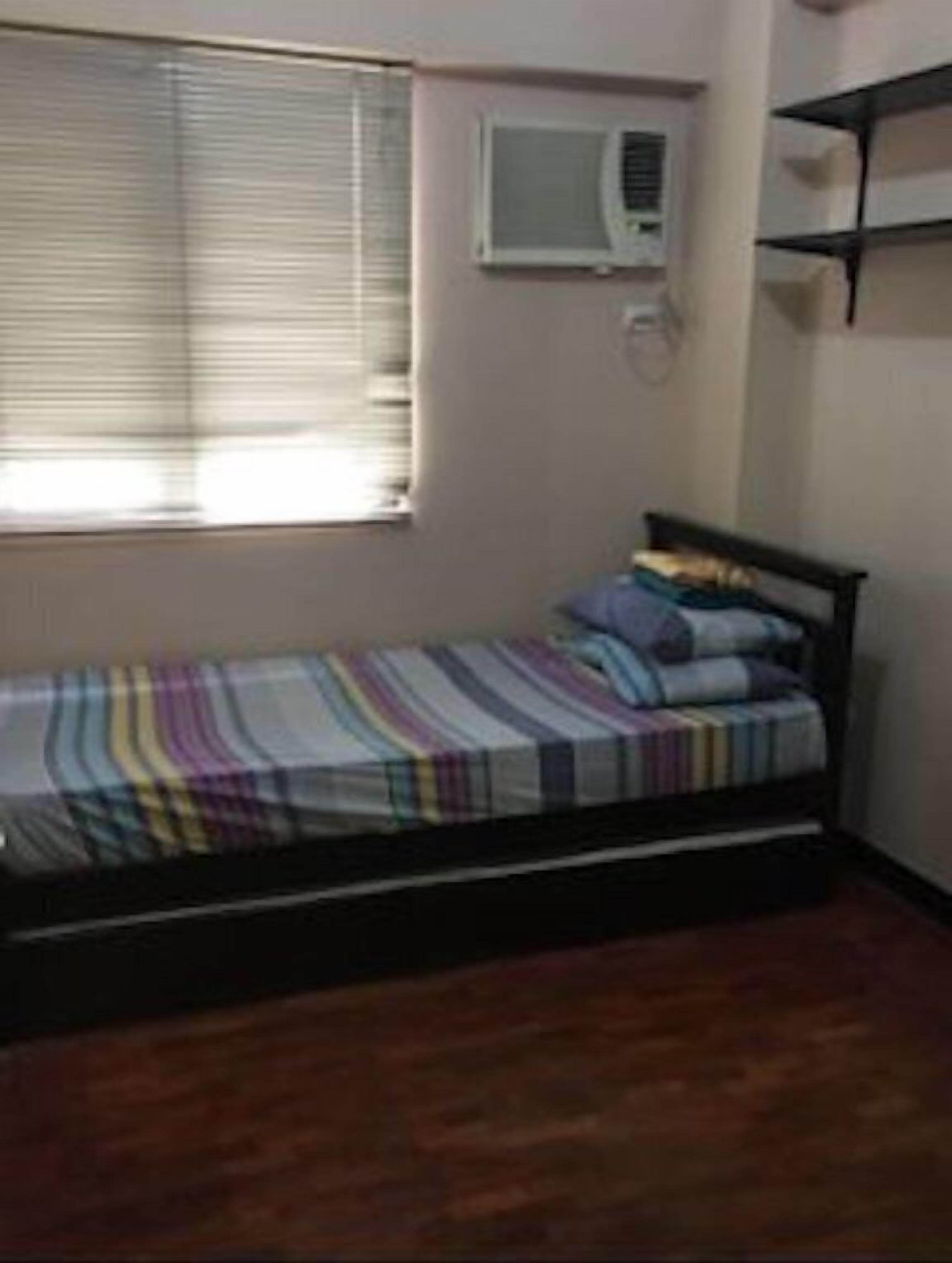 A small, single-bed room with a wooden floor, a window with blinds, and a wall-mounted air conditioning unit.