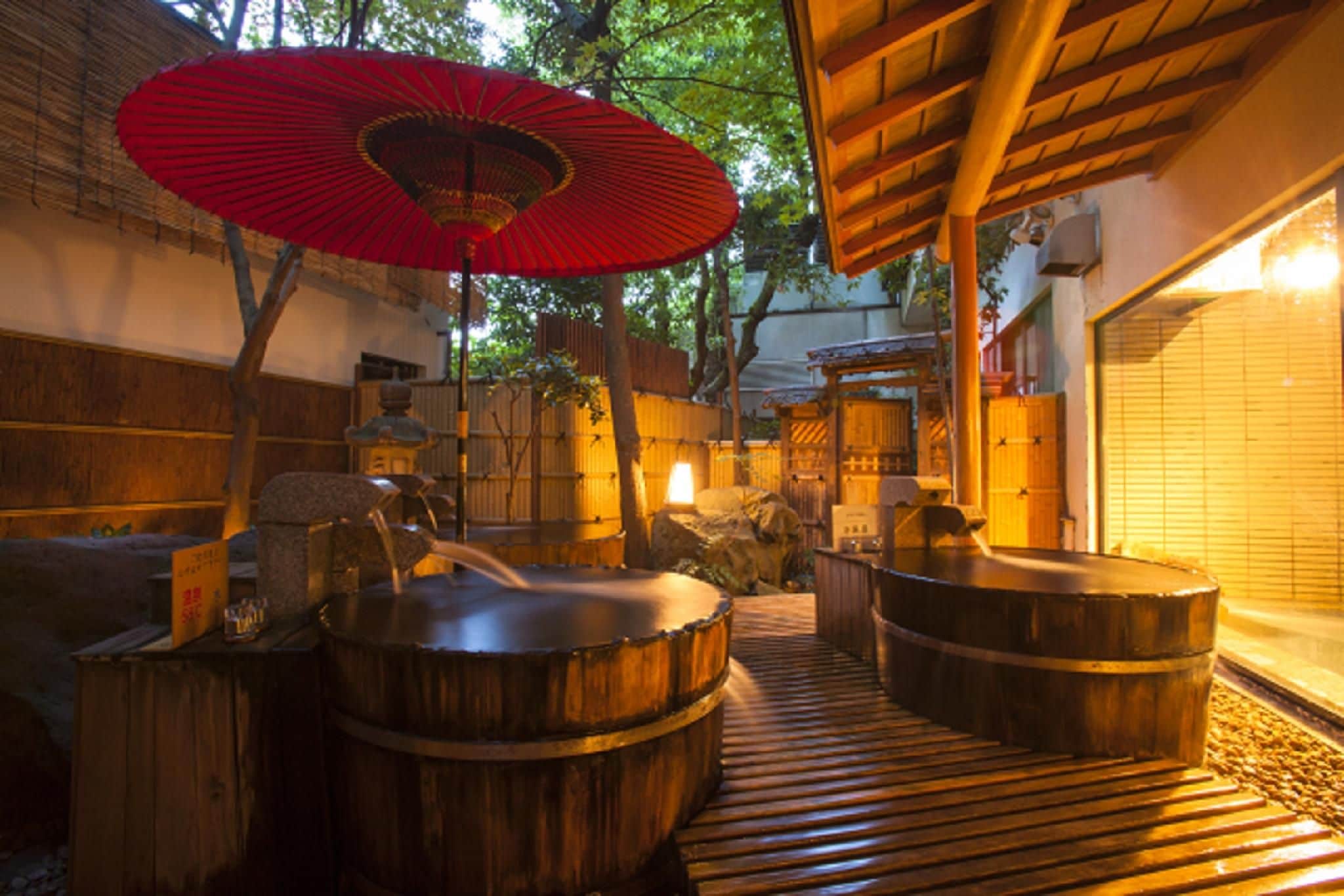Un onsen japonés tradicional con tinas de madera, un paraguas rojo y una estructura de madera.