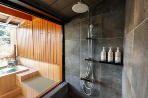 A modern bathroom with a wooden bench, a shower with a rain showerhead, and a shelf with toiletries. - Ryokufukaku (Toyooka)