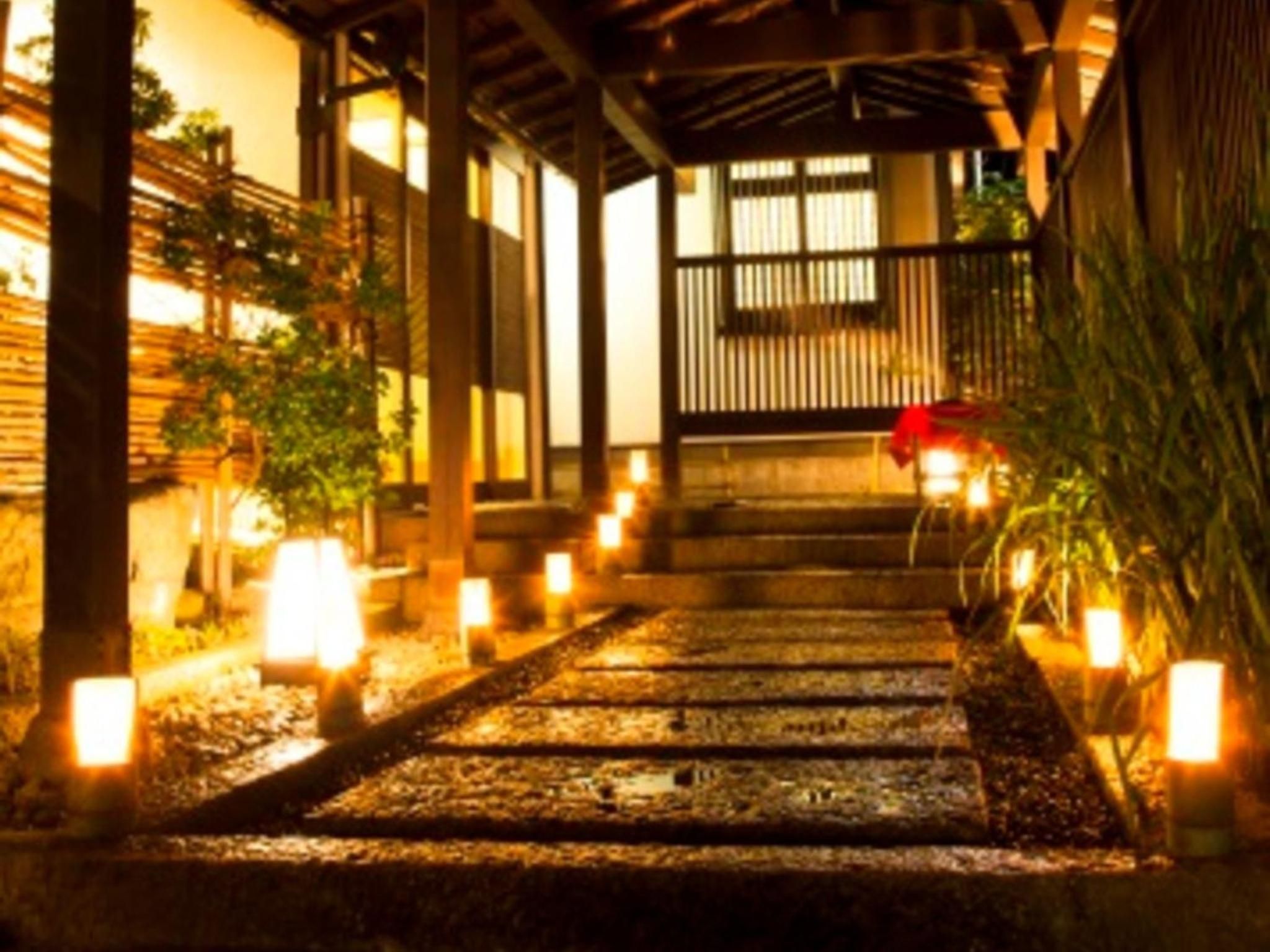 A pathway leading to a traditional wooden structure with a covered entrance, surrounded by plants and illuminated by candles.