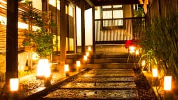 A pathway leading to a traditional wooden structure with a covered entrance, surrounded by plants and illuminated by candles.