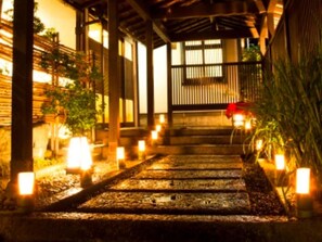 A pathway leading to a traditional wooden structure with a covered entrance, surrounded by plants and illuminated by candles. - Ryokufukaku (Toyooka)