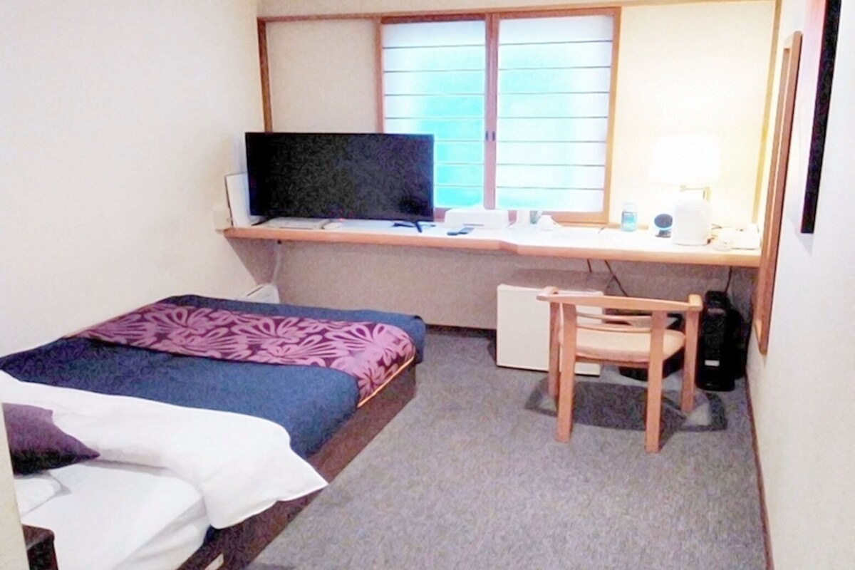 A small, neatly arranged room with a bed, a desk with a computer, and a chair.