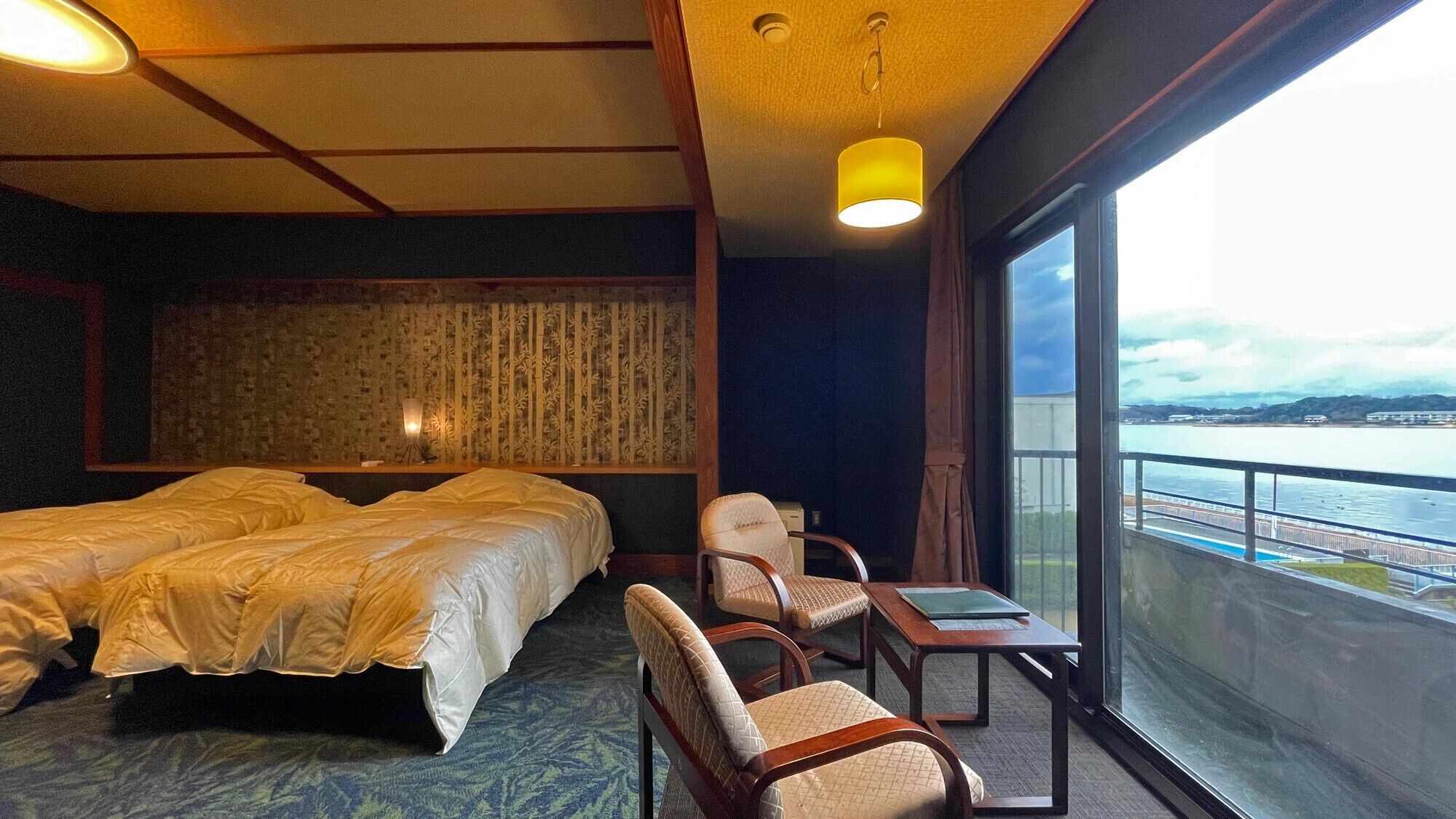Una habitación de hotel con cama, dos sillas, una mesita, una lámpara y vistas a un cuerpo de agua.