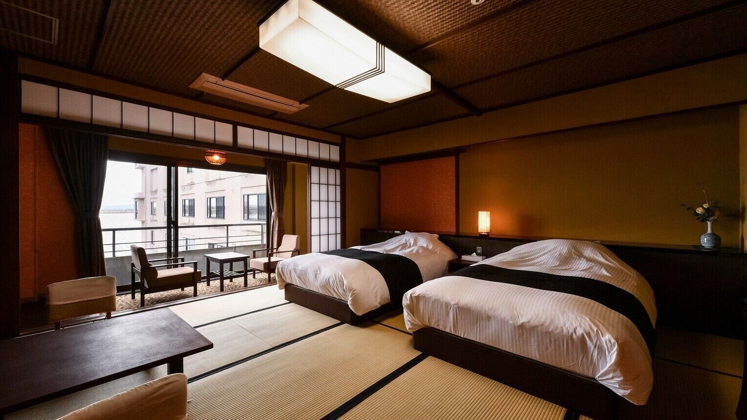 A hotel room with two beds, tatami mats, a balcony with seating, and a view of buildings.