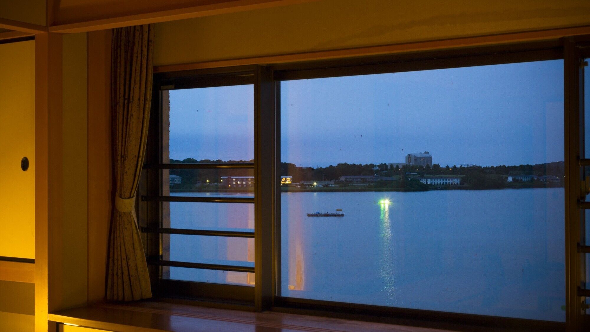 Una habitación con un ventanal grande con vistas a un cuerpo de agua al atardecer, una cortina y un suelo de madera.
