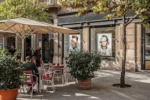An outdoor cafe with tables and chairs, a large umbrella, and a building with posters and a tree.
