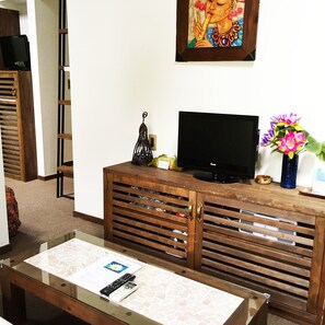 A living room with a television on a wooden cabinet, a coffee table with a remote control, a decorative candle, and a framed picture on the wall. - Estivant Club (Tsumagoi)