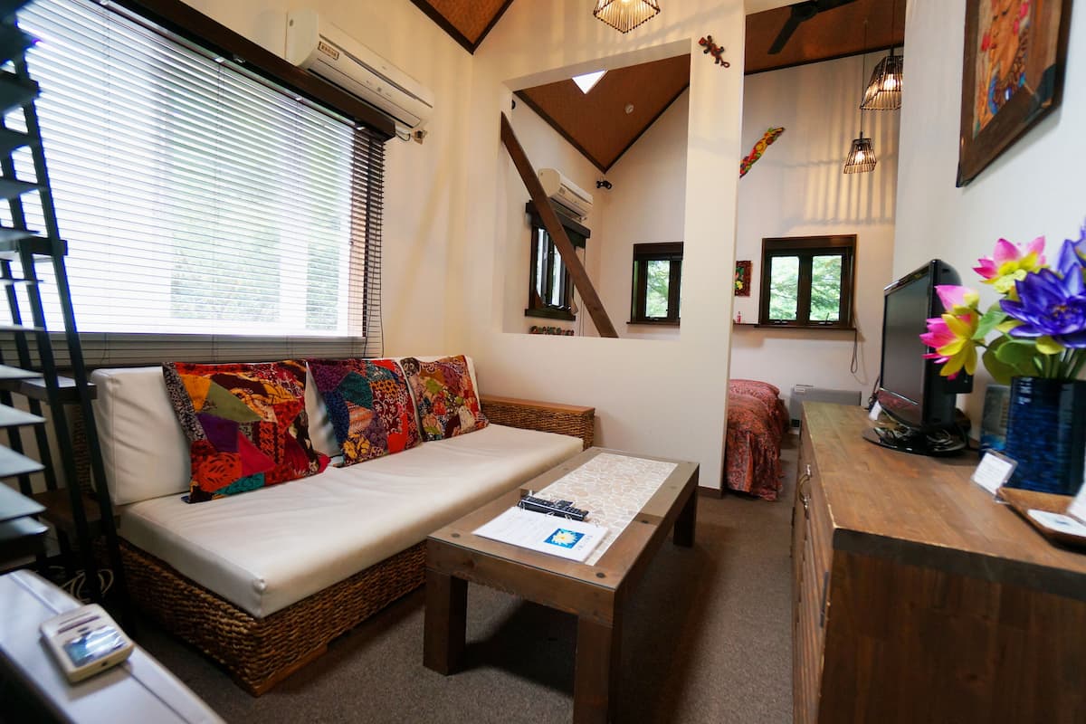 A cozy room with a sofa, a small table, and a wooden desk with a computer. There are windows with blinds, a ceiling fan, and a decorative wall hanging.