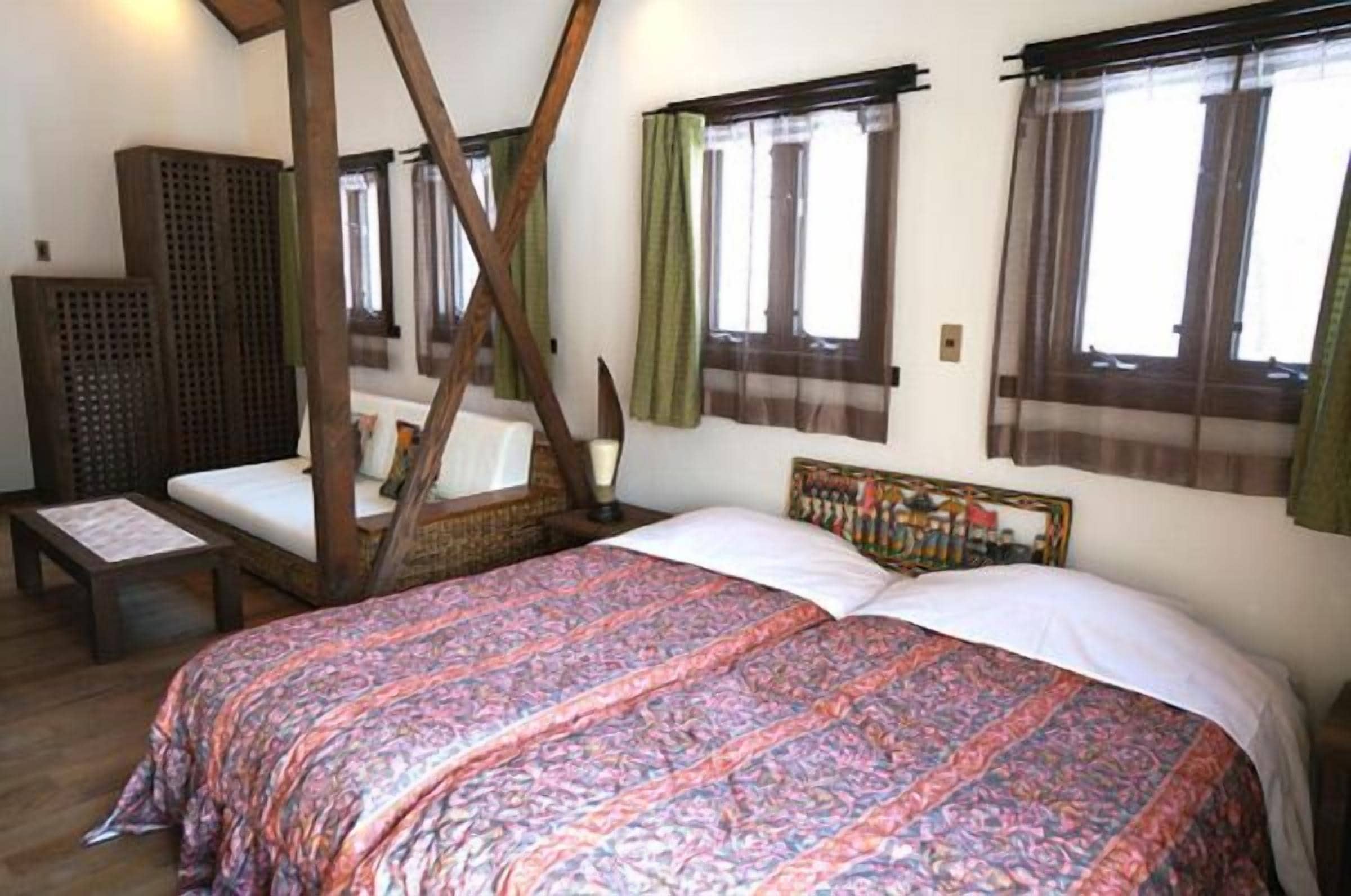 A room with a bed, a wooden chair, and a small table. There are windows with curtains and a wooden structure in the corner.
