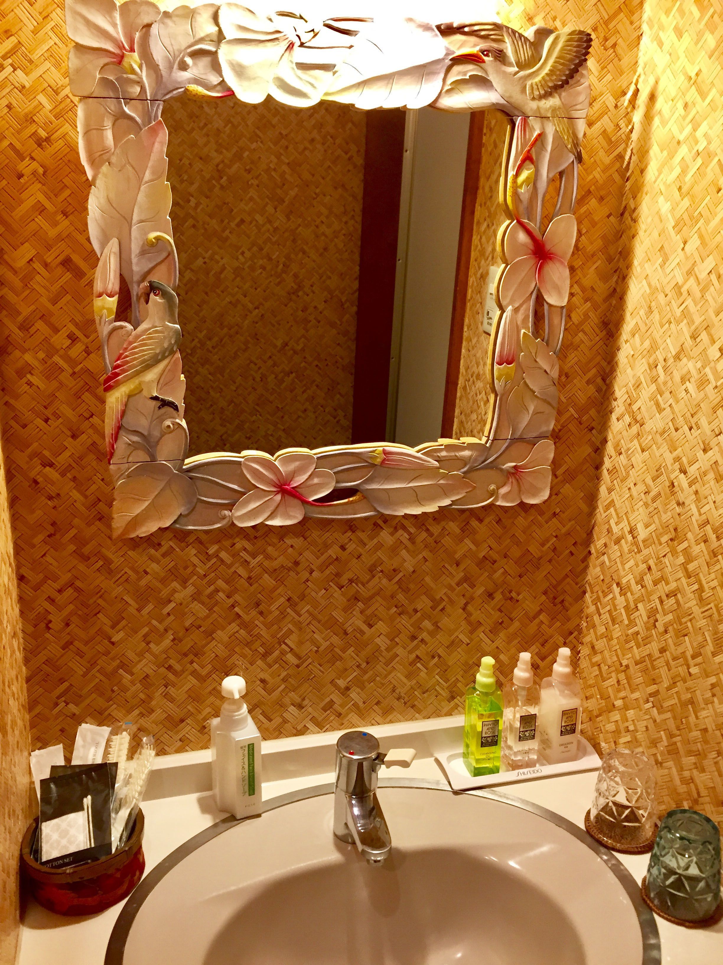 Un baño con un espejo de marco decorativo, un lavamanos, artículos de tocador y un panel de pared tejido.