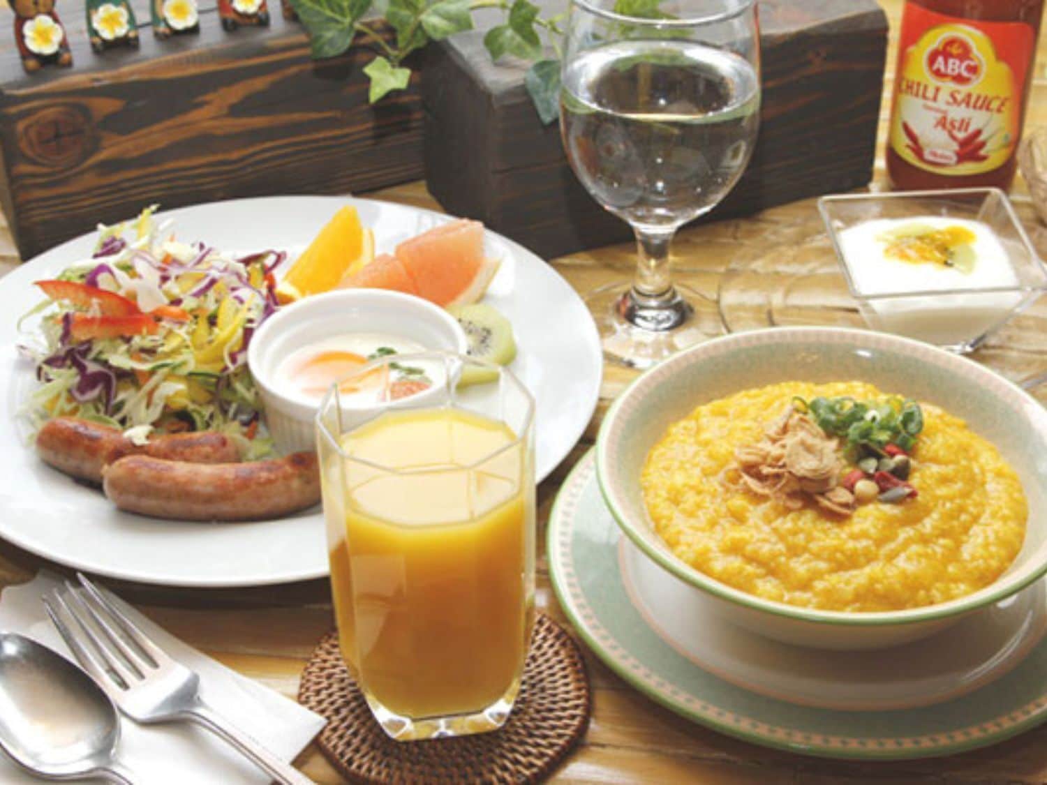 A meal with sausages, salad, and a bowl of porridge, along with a glass of orange juice and a bottle of chili sauce.