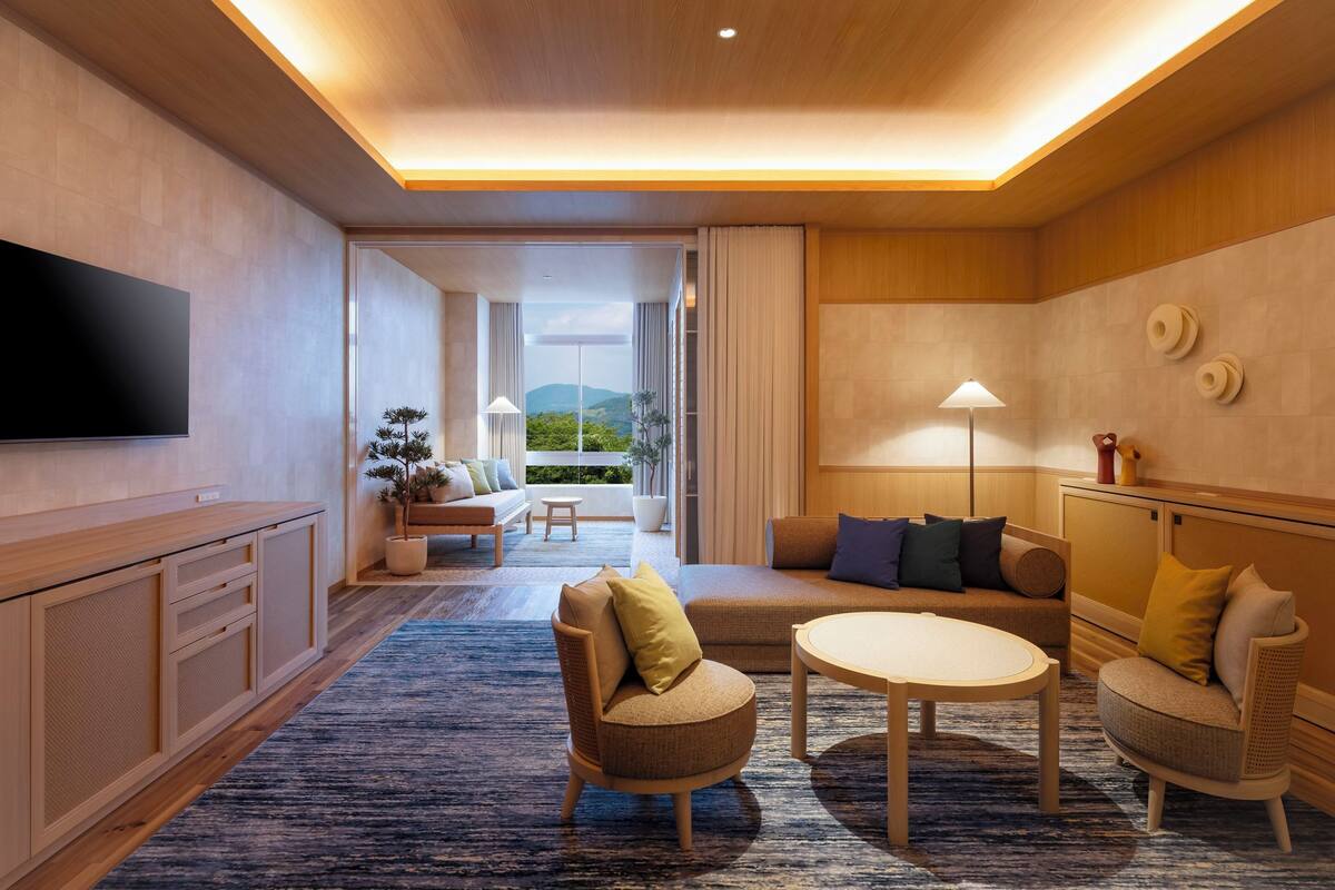 Ein modernes Hotelzimmer mit einem großen Flachbildfernseher, einer Couch, zwei Sesseln, einem kleinen Tisch und Blick ins Freie.