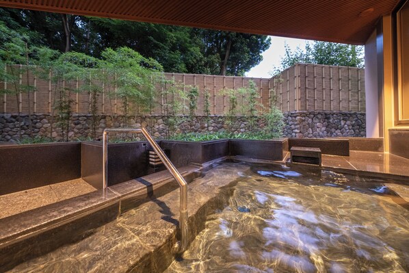 A hot spring area with stone steps leading into a clear pool, surrounded by a bamboo wall and a stone wall.