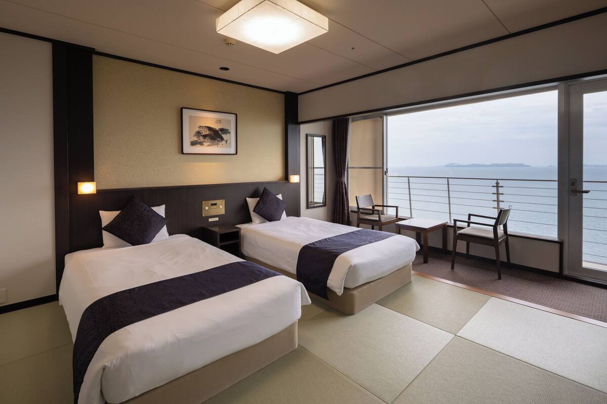 Ein Hotelzimmer mit zwei Betten, einem kleinen Tisch, Stühlen, einem großen Fenster mit Meerblick und einem gerahmten Bild an der Wand.