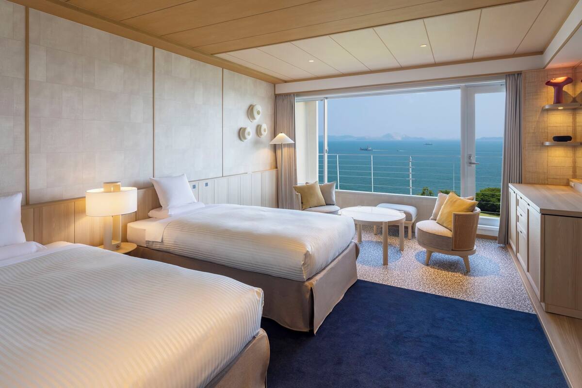 Ein Hotelzimmer mit zwei Betten, einer kleinen Sitzecke, Meerblick und einem Balkon mit Geländer.