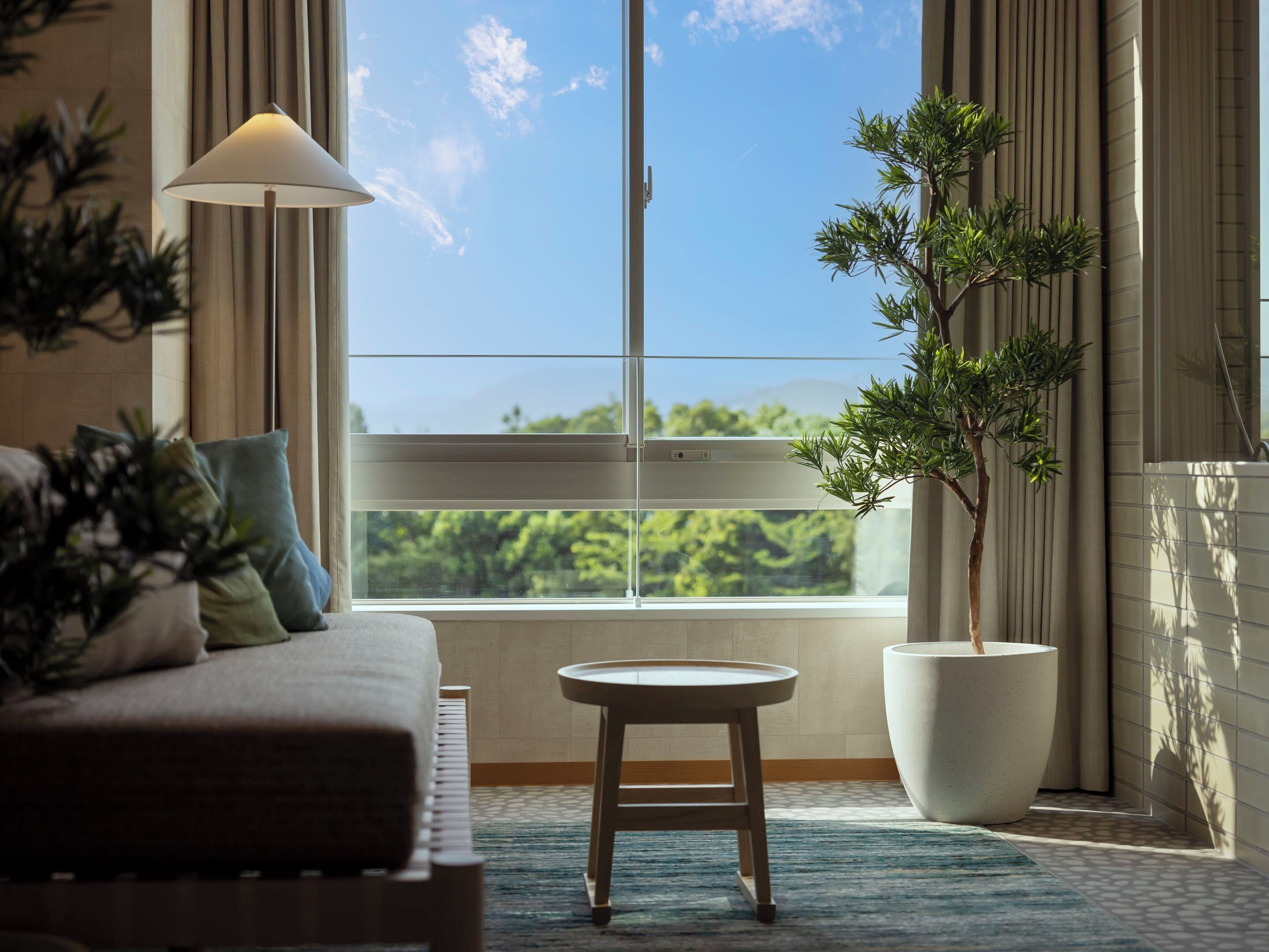 Un salón moderno con un sofá, una mesita redonda, una planta en maceta y un ventanal con vistas a zonas verdes.