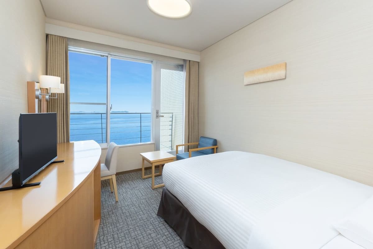 Una camera d'albergo con un letto, una scrivania con televisore, una sedia, un tavolino e una finestra vista mare.
