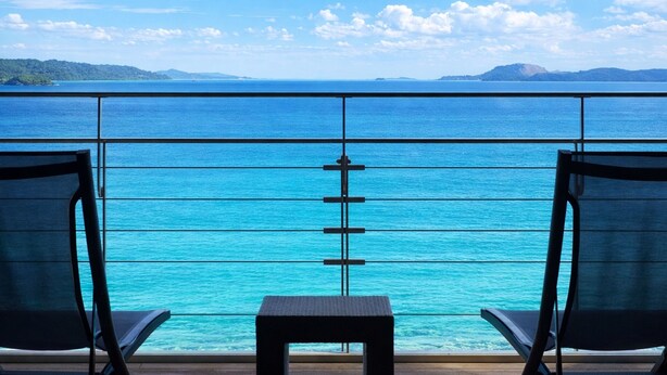 A balcony with two chairs and a small table, overlooking a turquoise sea and distant land under a clear sky.